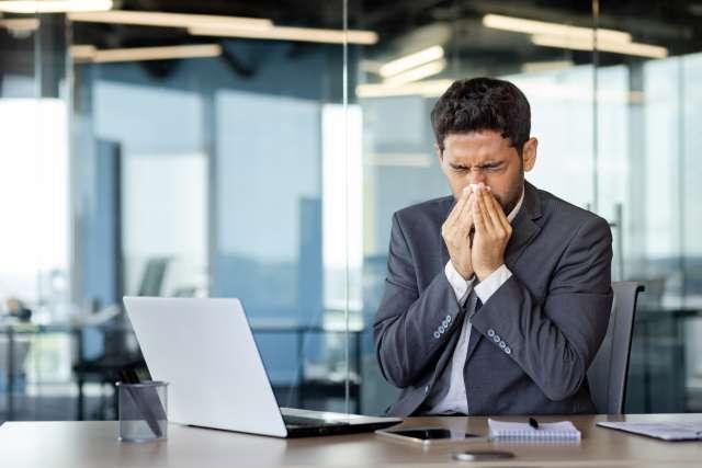 A man with allergies sneezes while at his work desk, covering his nose with a tissue.